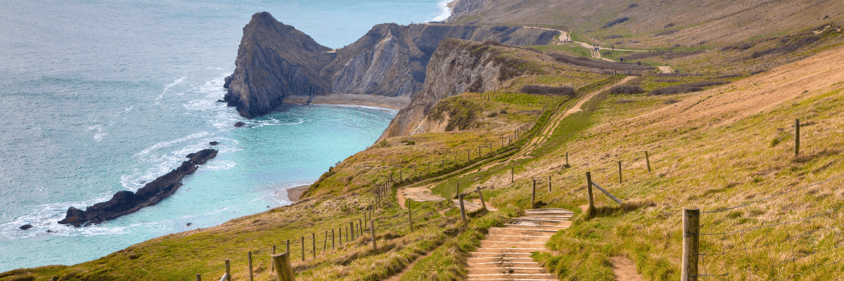 A scenic coastal hiking trail winding down a grassy hillside on a sunny day. The trail features wooden steps and is bordered by a rustic post-and-wire fence, overlooking dramatic limestone cliffs and a secluded sandy cove. The bright turquoise ocean below is dotted with a distinctive line of dark rocks jutting out of the water, capturing the rugged beauty of the Jurassic Coast.