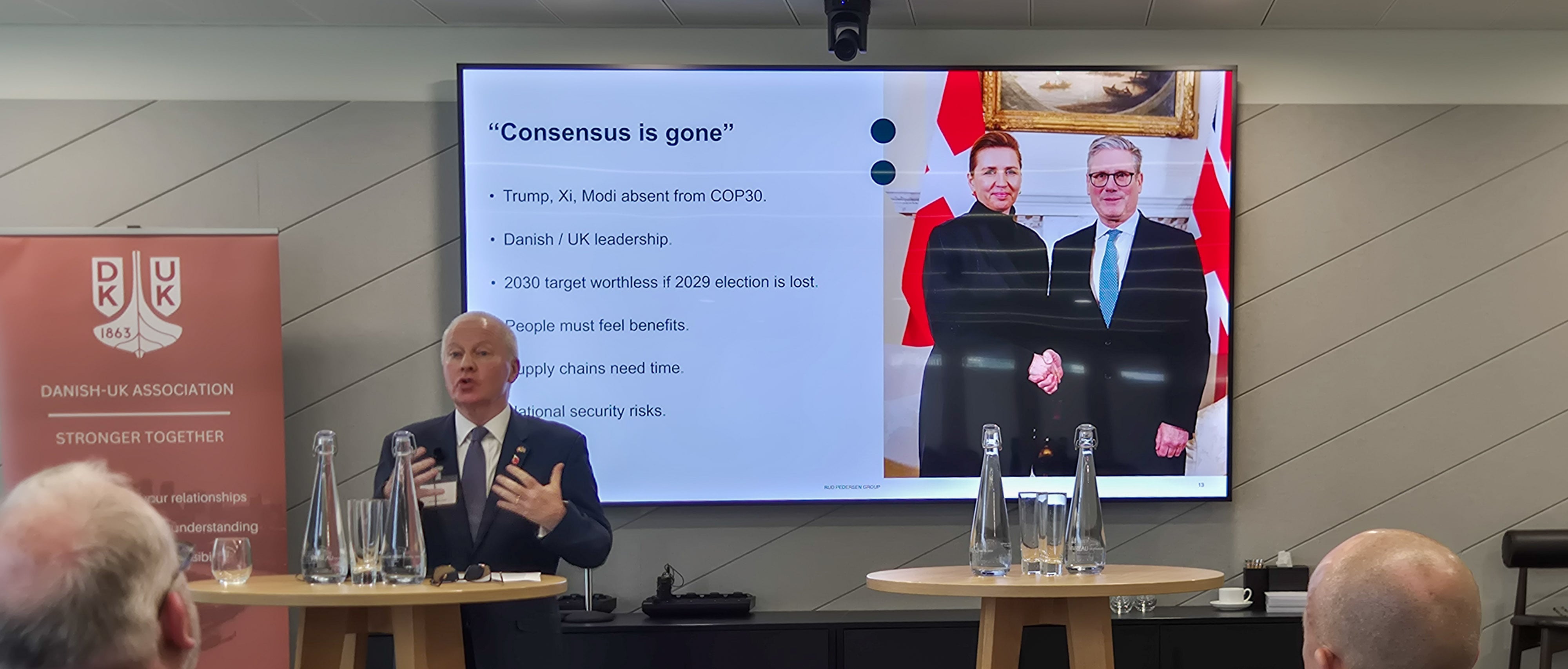 A speaker from the Rud Pedersen Group presents at the ReCharge 2025 event in London, standing beside a Danish‑UK Association banner. On the screen behind him, a slide titled “Consensus is gone” lists geopolitical and policy challenges, alongside a photo of Danish Prime Minister Mette Frederiksen shaking hands with UK Prime Minister Keir Starmer.